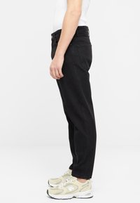 Jeans tapered negros de mezclilla, con una cintura de altura media y bolsillos laterales, combinados con zapatillas de color claro. Textura suave.