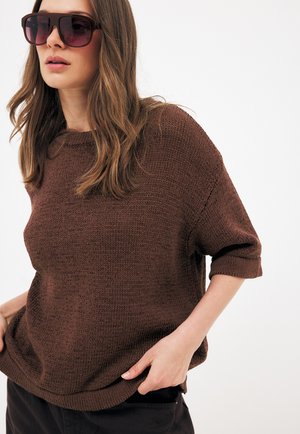 Kvinde iført oversized brun strikket sweater og store mørke solbriller, med løst bølget hår, poserer med den ene hånd i lommen mod hvid baggrund.
