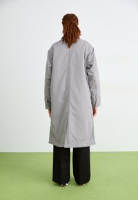 Cappotto lungo grigio con una vestibilità rilassata, caratterizzato da uno spacco posteriore e polsini con bottoni, abbinato a pantaloni neri a gamba larga e calzature decorate.