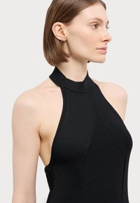 Musta ribbed-halter-kaula mekko, jossa on tiukka siluetti. Siinä on korkea kaulus ja teksturoitu kangas. Asusteena pyöreät korvakorut.