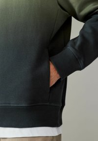 Tamnozeleni dukserica sa rebrastim rubovima i manžetama, koja ima bočnu džepu. Materijal se čini glatkim i ugodne teksture.