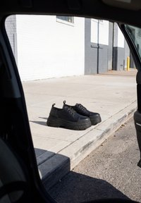 Coppia di scarpe nere con suola platform spessa posate su un marciapiede di cemento accanto a un cordolo, viste dall'interno di una porta aperta di un'auto.