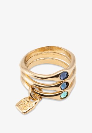 Anillos apilados de tono dorado con piedras azules y moradas, llevados en una mano parcialmente visible contra un fondo neutral. Acabado suave y pulido.