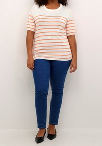 White and orange striped knit top with short sleeves, paired with blue skinny jeans and black flats, all set against a plain background.