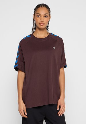 Femme aux cheveux tressés portant un T-shirt ample marron foncé avec des rayures bleues en chevrons sur les manches et un petit logo blanc sur la poitrine.