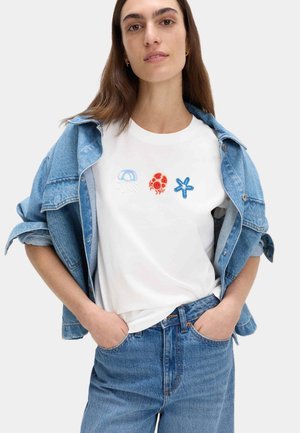 Vrouw draagt een wit T-shirt met borduursels van kwal, krab en zeester, gecombineerd met een spijkerjasje en blauwe jeans, handen in zakken.