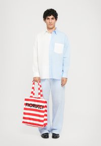 Ung man klädd i en halvt vit, halvt blå skjorta och randiga byxor, som håller i en röd och vit randig Fiorucci-axelväska.