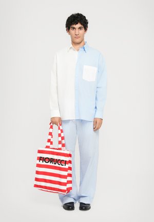 Ung mann iført en halv-hvit, halv-blå skjorte og stripete bukser, holder en rød og hvitstripet Fiorucci totebag.