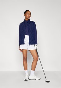 Mornarica, podstavljena jakna s visokim ovratnikom, bijele golf šortse i bijele tenisice. Drži golf palicu i nosi bijelu rukavicu. Glatka tekstura.