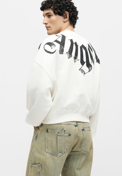 Άνδρας με κοντά σγουρά μαλλιά φοράει λευκό φαρδύ πουλόβερ με μαύρο γοτθικό κείμενο «Ang» στην πλάτη, σε συνδυασμό με φαρδιά ξεβαμμένα μπεζ τζιν.