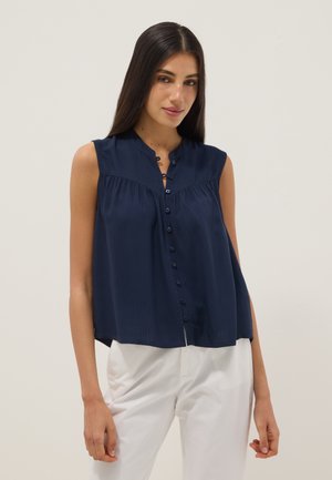 Vrouw draagt een mouwloze marineblauwe blouse met knopen en witte broek, staand tegen een effen lichte achtergrond.