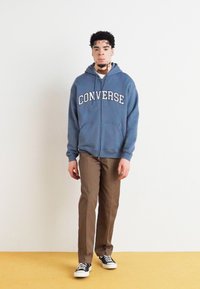 Felpa blu con zip e la scritta "CONVERSE" in bianco, abbinata a pantaloni marroni e sneaker bianche e nere. Sfondo semplice.