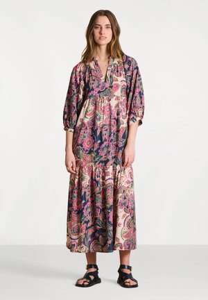 Femme portant une longue robe fleurie ample aux motifs roses, verts et bleu marine, associée à des sandales noires à brides, debout sur un fond blanc uni.