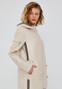 Beige leather jacket with a hood, featuring silver hardware, side pockets, and black vertical stripes along the sides. Smooth texture.