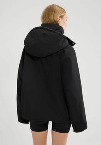 Czarna kurtka z kapturem o luźnym kroju, z wysokim kołnierzem i gładką fakturą. Widok z tyłu podkreśla minimalistyczny design i brak widocznych zapięć.