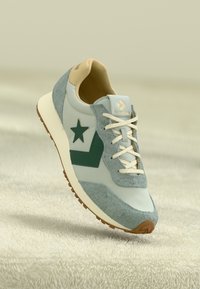 Light blue sneakers with gray suede accents, green star and chevron design, cream laces, tan inner lining, and textured brown outsole.