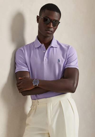 Πολύχρωμο πουκάμισο πόλο με κοντά μανίκια σε αποχρώσεις λεβάντας, με μικρό λογότυπο, συνδυασμένο με κρεμ πλισέ παντελόνια. Συμπληρώνεται με ρολόι με μπλε καντράν και γυαλιά ηλίου.