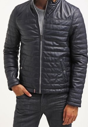 Mann trägt schwarze gesteppten Reißverschlussjacke über grauem Shirt mit schwarzen Hosen, linke Hand in der Tasche, rechte Hand entspannt an der Seite.