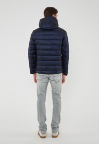 Uomo che indossa un giubbotto imbottito blu navy con cappuccio, jeans grigi chiari e sneaker bianche, in piedi di spalle su uno sfondo semplice.