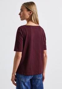 Bordeaux kleurig t-shirt met korte mouwen, brede halslijn, ontspannen pasvorm en een zachte textuur, gecombineerd met blauwe denim broek.