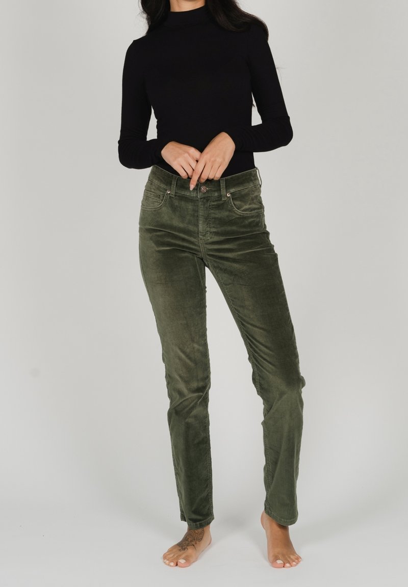 Femme en haut noir à manches longues portant un pantalon en velours côtelé vert olive, debout pieds nus sur un fond clair uni.