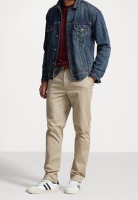 Muž v modré denimové bundě, vínovém tričku, béžových kalhotách s opaskem a bílých teniskách s modrými pruhy, stojící s rukama v kapsách.