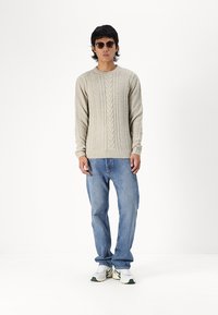 Suéter de punto beige con mangas largas, combinado con jeans de color azul claro y zapatillas blancas con detalles en verde. Unas gafas de sol marrones completan el look.