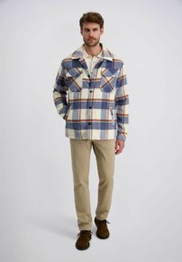 Man in een blauw, bruin en witte geruite jas over een beige shirt met kaki broek en bruine schoenen, staande tegen een witte achtergrond.