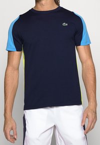 Tmavě modré sportovní tričko s modrými rukávy a žlutými akcenty, vyrobené z hladké, pružné látky; na hrudi je malé zelené logo.