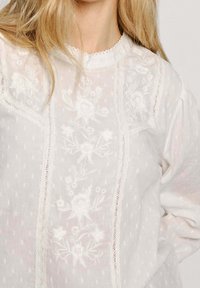 Weiße Bluse mit floralem Stickerei und Spitzenbesatz an der Vorderseite, mit einem gewellten hohen Kragen und langen Ärmeln.