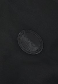 Tissu noir avec une texture lisse, présentant un patch en caoutchouc noir, embossé avec "Barbour" et entouré d'une bordure en fil noir.
