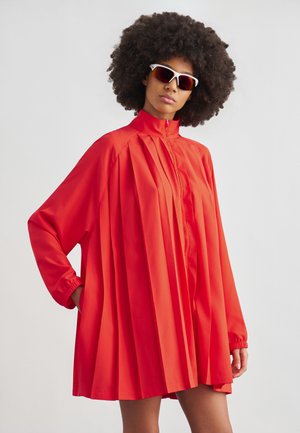 Donna con capelli ricci e voluminosi che indossa un vestito plissettato rosso con cerniera frontale e occhiali da sole sportivi bianchi, in piedi davanti a uno sfondo semplice.