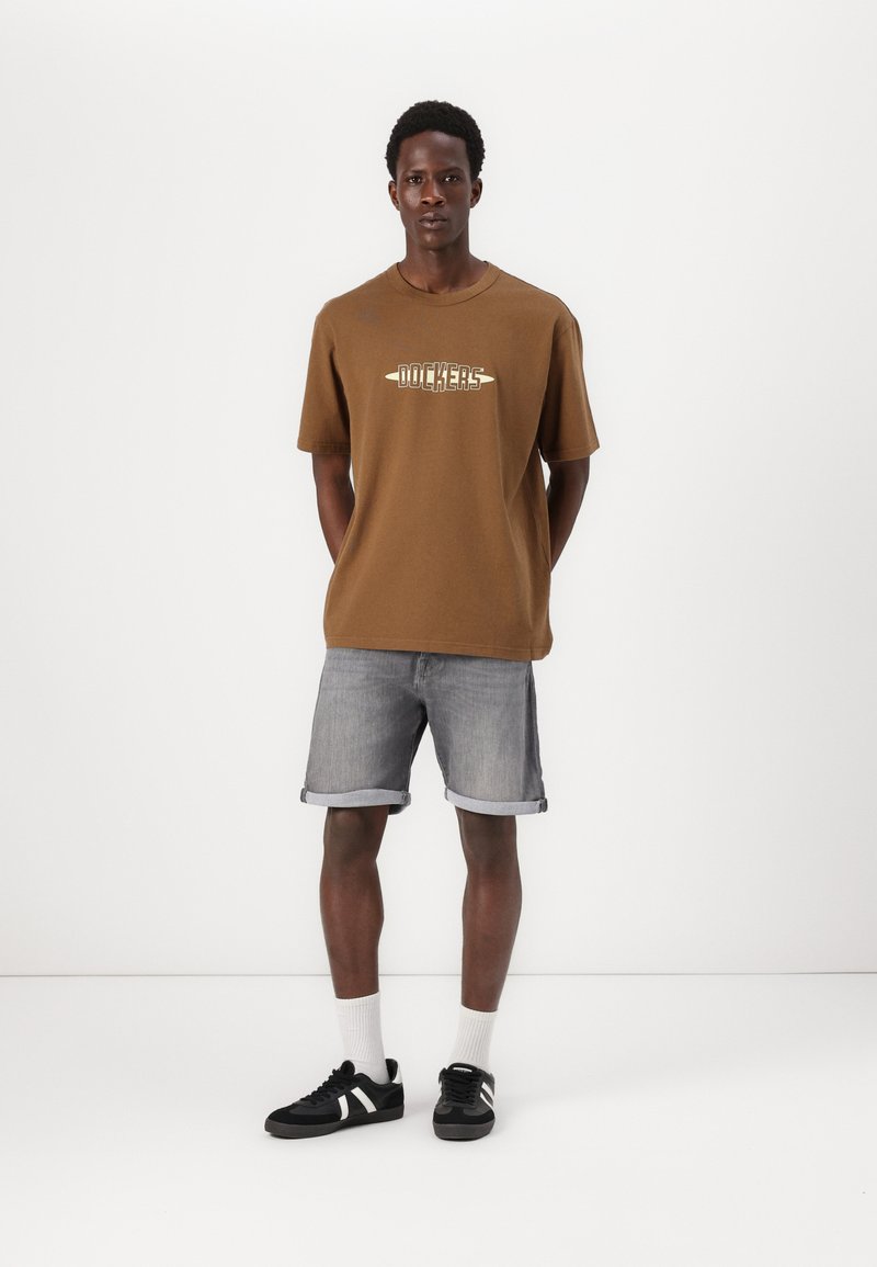 T-shirt en coton marron avec le logo "Dockers", associé à un short en denim gris clair, des baskets noires avec des rayures blanches, et des chaussettes blanches.