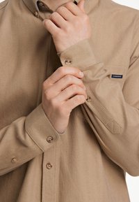 Beige shirt met een vissengraatpatroon, met een button-down kraag, lange mouwen en houten knopen, voorzien van een klein geborduurd merkembleem.