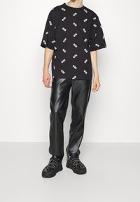 Camiseta negra con logotipos blancos, corte holgado, mangas cortas; combinada con pantalones de cuero negro ceñidos, que presentan una textura suave. Zapatillas negras con acentos.