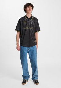 Μαύρο πόλο μπλουζάκι με κίτρινες λεπτομέρειες και λογότυπο, κοντά μανίκια, ελαφρύ υλικό, συνδυασμένο με άνετους μπλε τζιν και μαύρα παπούτσια.