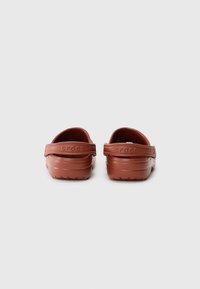 Brown rubber clogs with a rounded back, heel strap, and textured sole. "crocs" logo embossed on the strap.