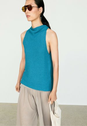 Frau mit dunkel zurückgebundenem Haar, trägt braune Sonnenbrille, ein blaues ärmelloses Stricktop, beige weite Hosen, hält eine weiße Tasche.