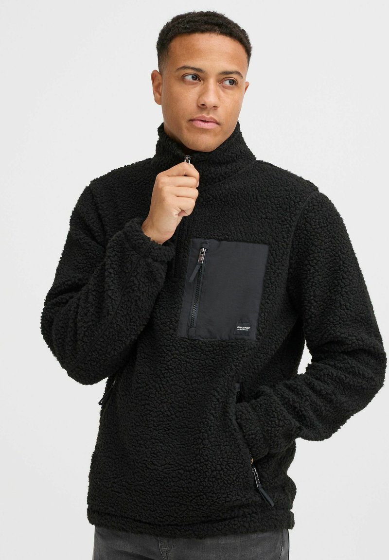 Black fleece pullover with a high collar, zippered chest pocket, and side pockets. Textured fabric and black panel detail on the chest.