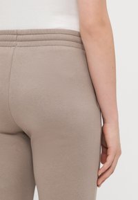 Pantaloni de trening bej cu o textură moale și un tiv elastic, având un detaliu discret cu dungi aproape de talie și o buzunar lateral.
