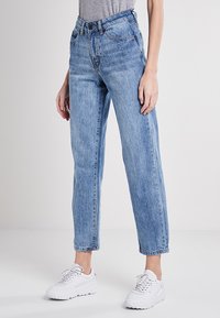 Jeans de tiro alto azul claro con un ajuste relajado, con un lavado desgastado, diseño de cinco bolsillos y costuras en contraste. Combinados con zapatillas blancas.