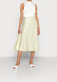 Žena v bílém bezrukávovém roláku, světle zelené midi sukni s jemným vzorem, bílých sandálech na podpatku a držící odpovídající zelenou kabelku.