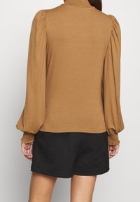 Braunes Langarmoberteil mit Stehkragen, puffigen Ärmeln und gerafften Details an den Manschetten. Kombiniert mit schwarzen Shorts. Glatte Textur.