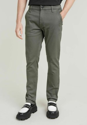 Olive green slim-fit trousers with a flat front, two front pockets, and a button closure, paired with black and white chunky shoes.