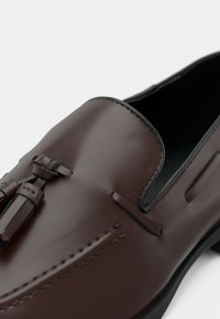 Mocassins en cuir marron présentant une finition lisse, des lacets décoratifs et une silhouette élégante avec des accents noirs contrastants le long de la semelle.