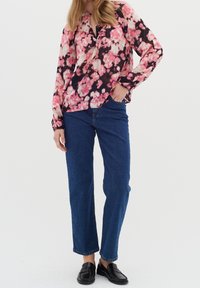 Persona che indossa una blusa a maniche lunghe floreale rosa e nera, jeans blu e mocassini neri, che sta in piedi con una mano in tasca.
