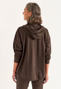 Brun hættebluse, let materiale, afslappet pasform, lange ærmer, sideslids, synlige sømdetaljer ved hætte og manchetter.
