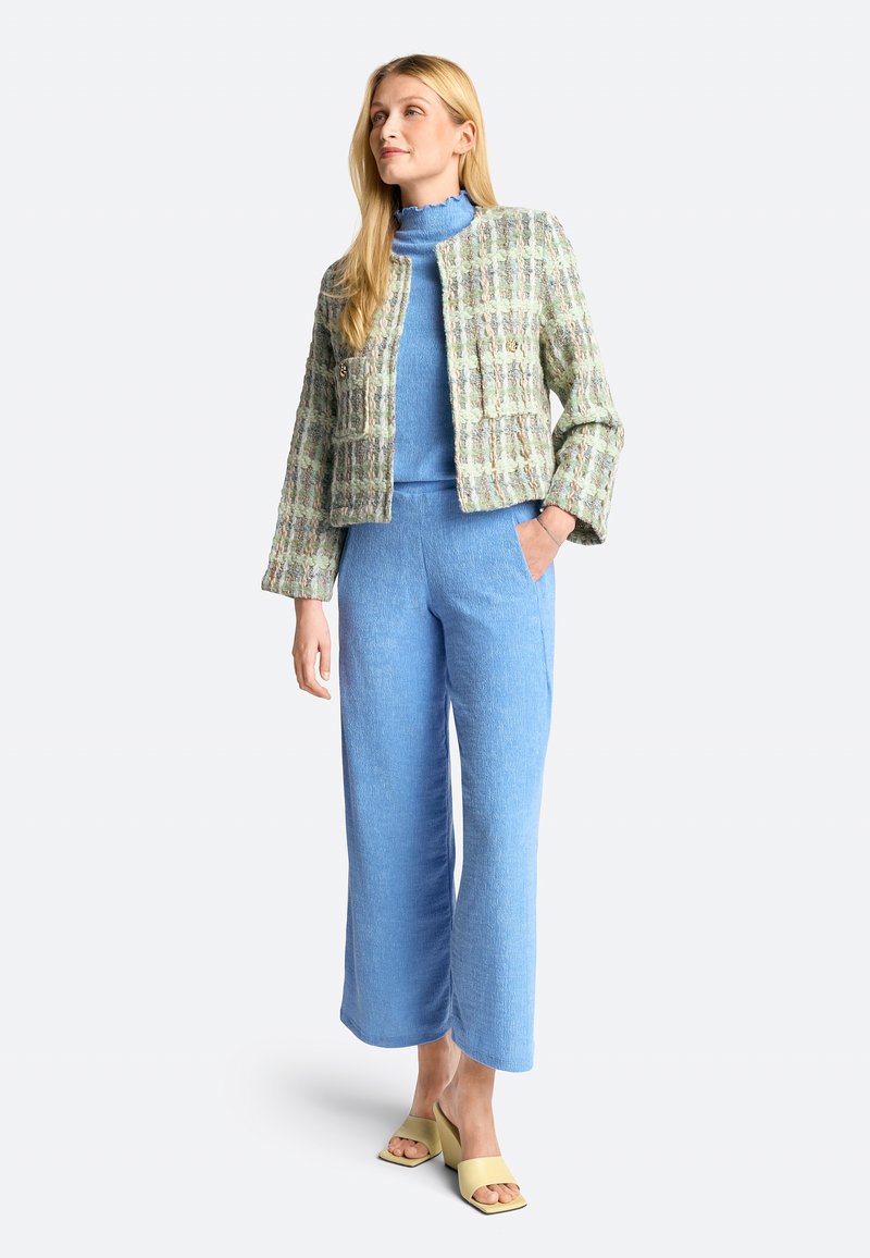 Hellgrüner und weiß gemusterter Tweed-Jacke mit zwei Taschen, kombiniert mit einem blauen Rollkragenpullover und weit geschnittenen blauen Hosen. Gelbe Schlüpfsandalen.