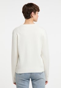 Pull beige clair en maille douce ; possède des manches longues, un col rond et des poignets et un ourlet côtelés, présenté de dos.