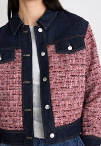 Chaqueta de mezclilla azul con un patrón tejido en tonos rosa y rojo, que cuenta con botones a presión plateados y un cuello con solapa.